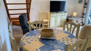 Un coin repas avec une table ronde recouverte d’une nappe à motifs bleus et jaunes, entourée de chaises en bois. Un bol en bois est posé sur la table.