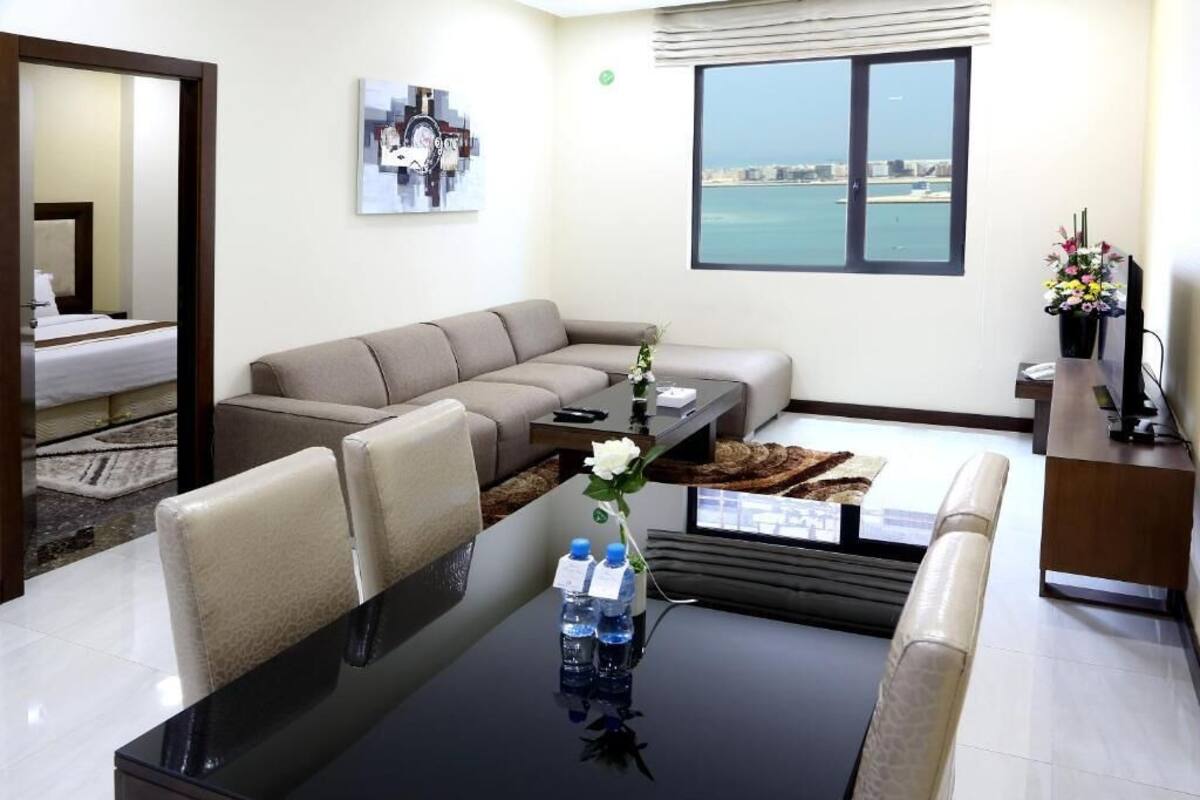 A modern living room with a sofa, coffee table, and chairs. A large window offers a view of the sea and cityscape.
