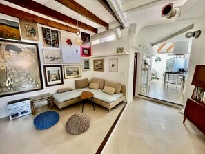 Uma sala de estar moderna com um sofá seccional, uma mesa de centro e uma grande moldura na parede. Uma cozinha é visível através de uma porta aberta.