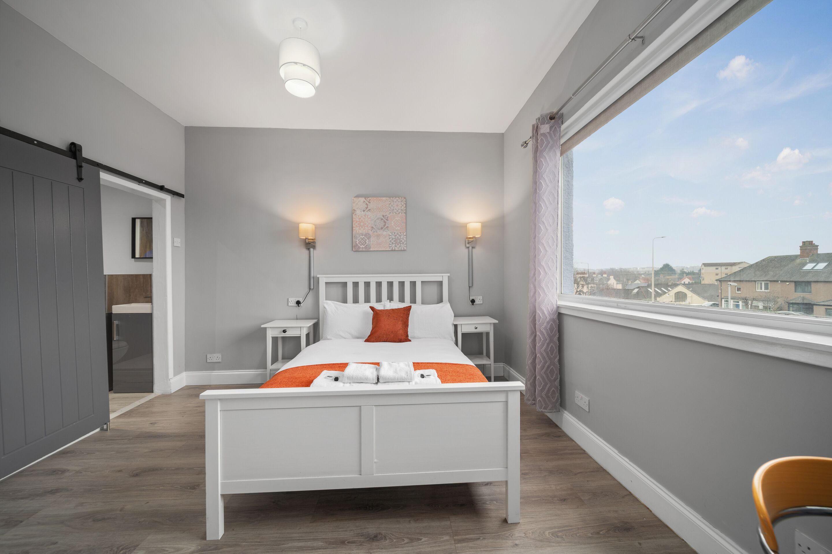 A modern bedroom with a white bed, two bedside lamps, a view of the city through a large window, and a cityscape visible outside.