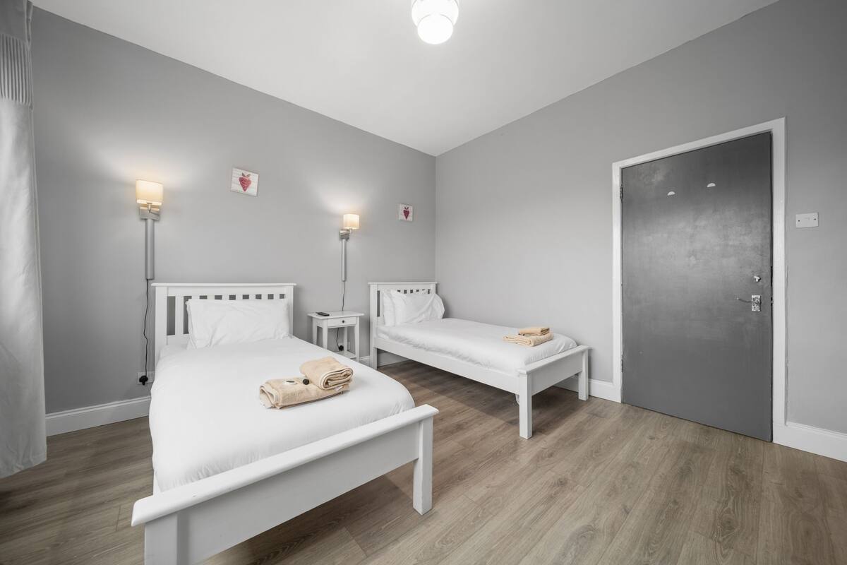 A room with two single beds, a bedside table with a lamp, a door, and a wall-mounted light fixture.