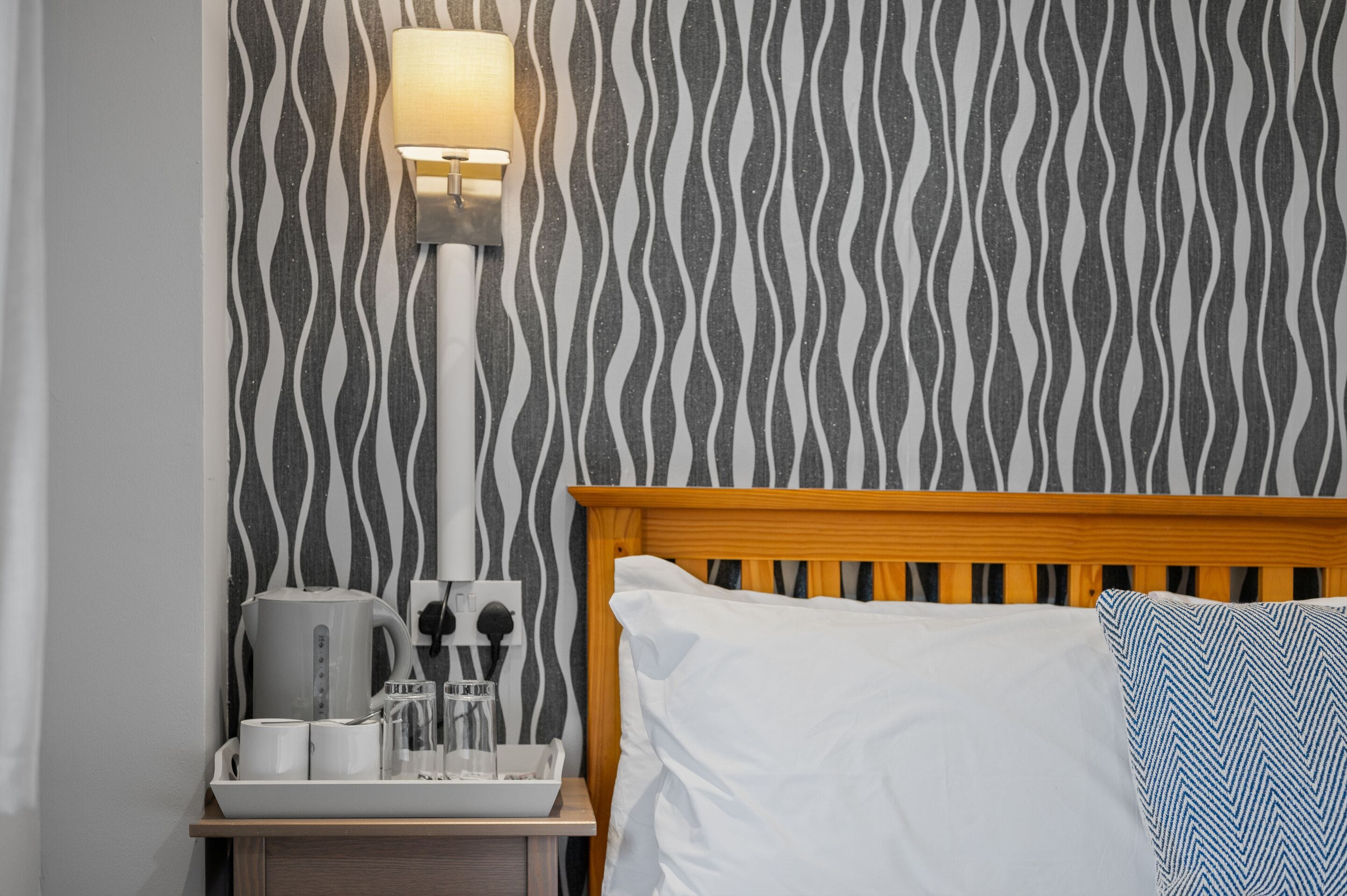 A hotel room with a zebra-striped wallpaper, a wooden headboard, a bedside table with a kettle, cups, and glasses, and a lamp.