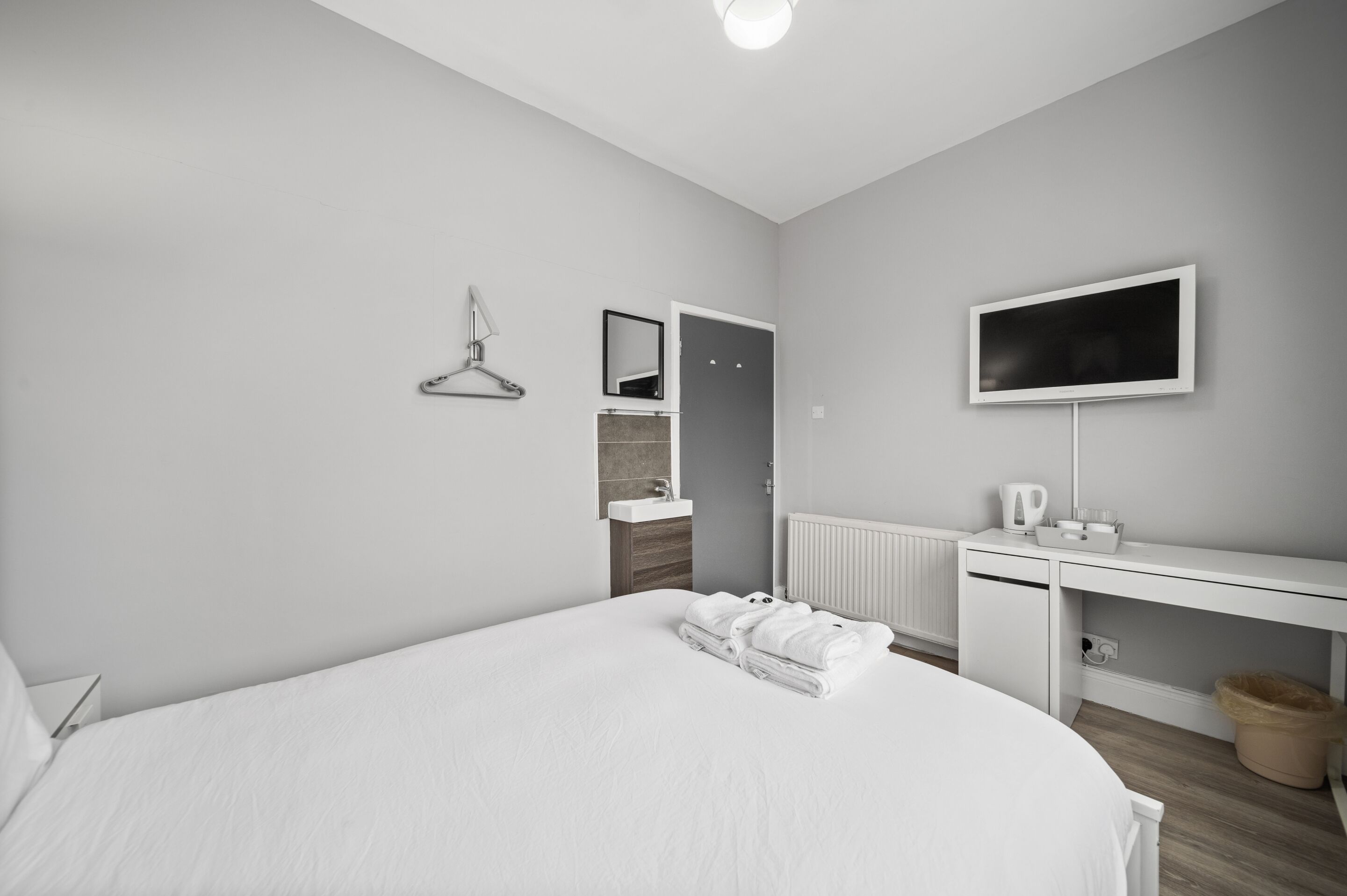 A modern hotel room with a large bed, a white desk with a flat-screen TV, a mirror, and a hanging towel rack.
