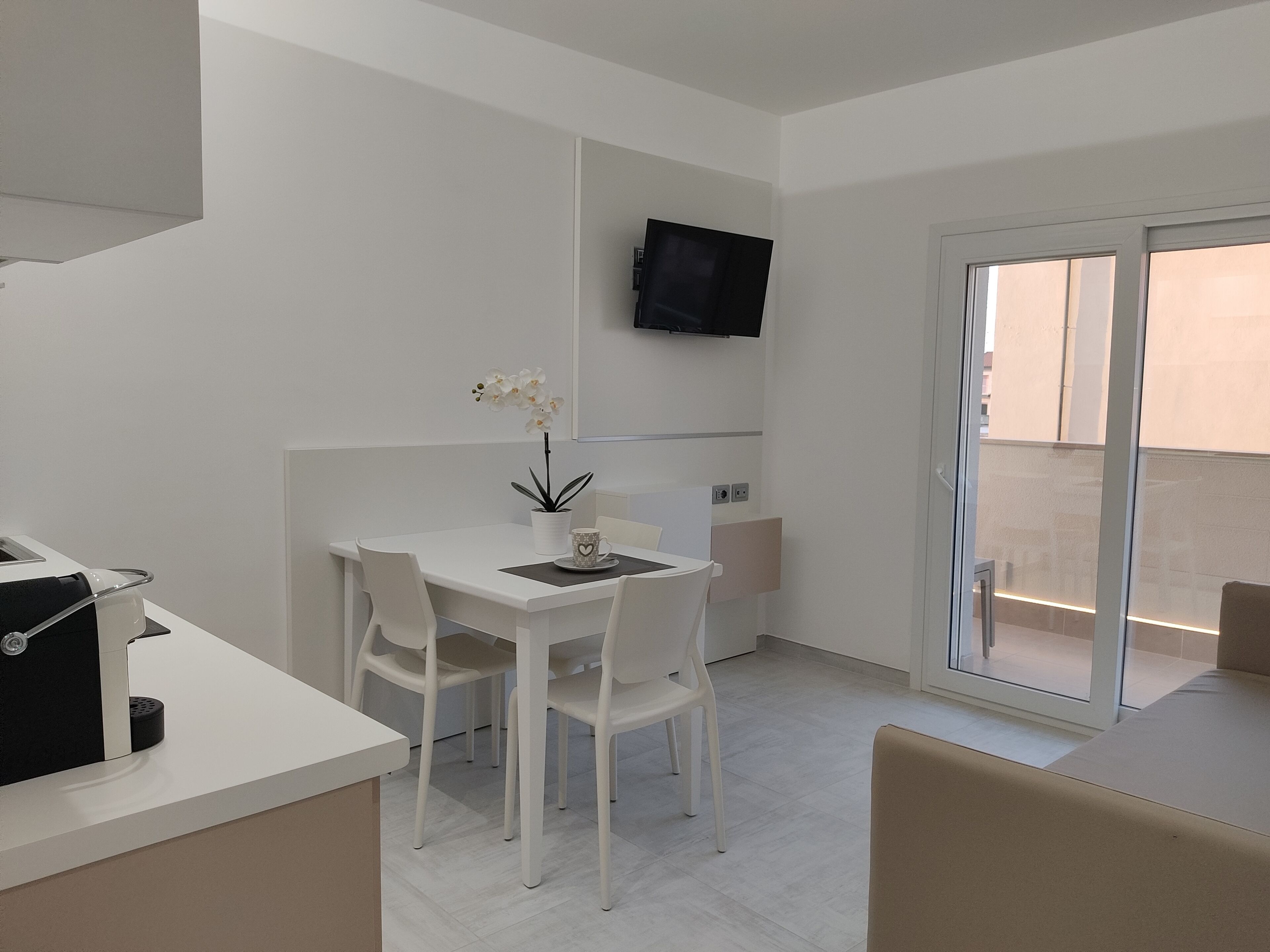 A modern, minimalist room with a white table and chairs, a small potted plant, a flat-screen TV mounted on the wall, and a sliding glass door leading to an outdoor area.
