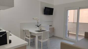 A modern, minimalist room with a white table and chairs, a small potted plant, a flat-screen TV mounted on the wall, and a sliding glass door leading to an outdoor area.