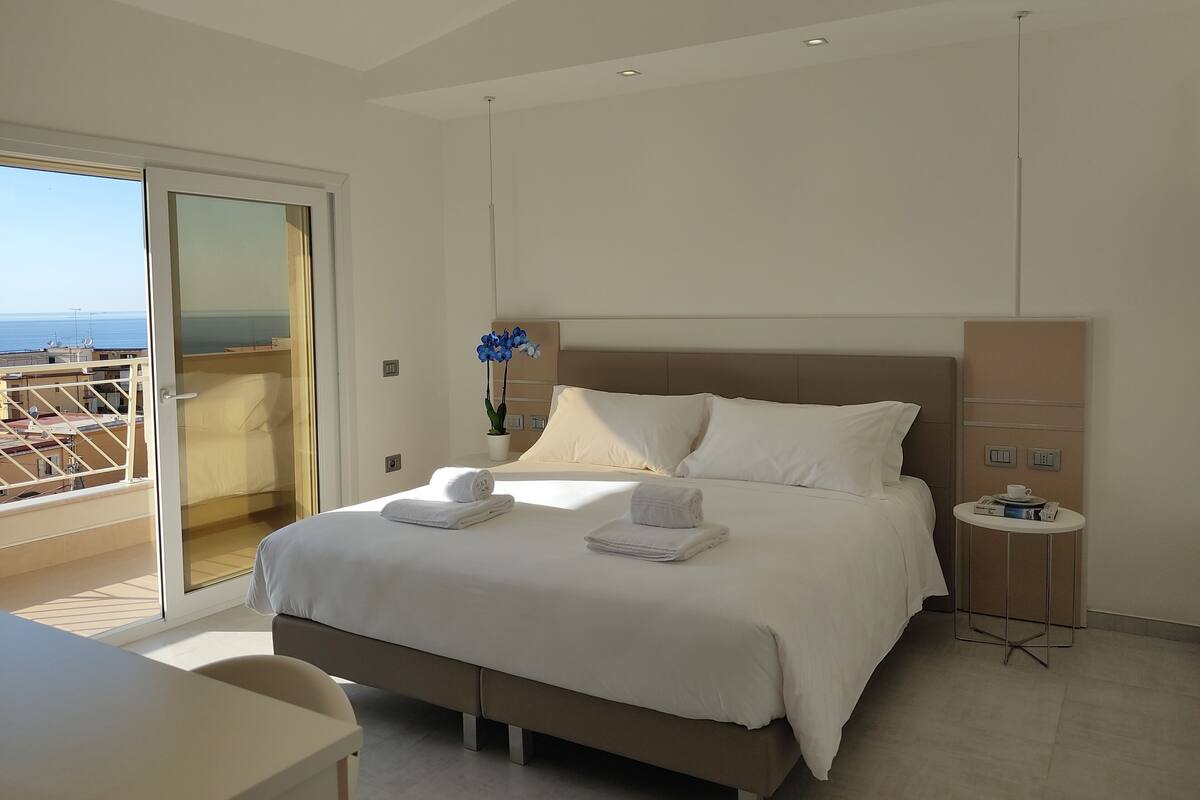 A modern hotel room with a large bed, a small bedside table, a view of the sea through a sliding door, and a potted plant.