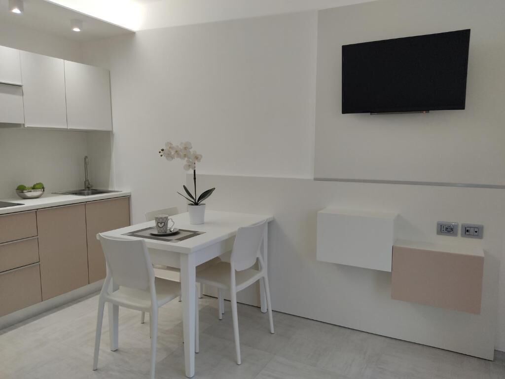 A modern kitchen with white cabinetry, a dining table with chairs, a potted orchid, and a mounted flat-screen TV.