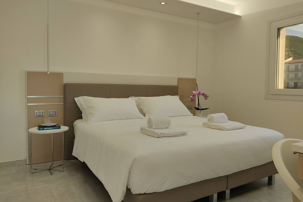 A modern hotel room with a large bed, a bedside table with a glass, a small table with a book, and a window with a view of buildings.