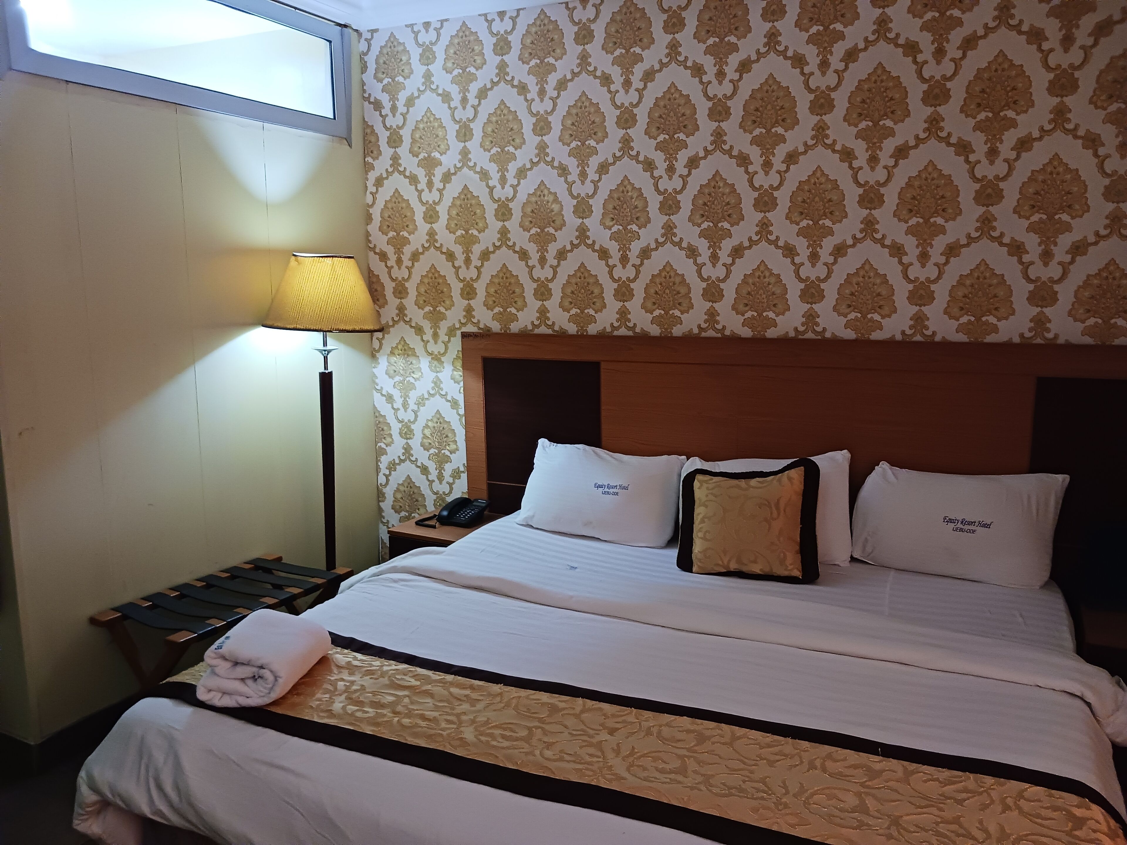 A hotel room with a large bed, a bedside table with a lamp, patterned wallpaper, and a headboard.