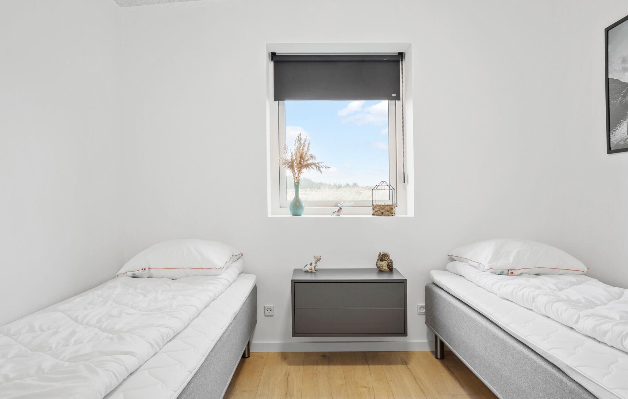 Une petite chambre moderne avec deux lits simples, une petite table de chevet, une fenêtre donnant sur un champ et un tableau encadré au mur.