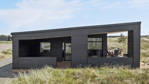 Une cabane moderne et minimaliste, dotée de grandes fenêtres et d’un toit plat, située dans une zone herbeuse sous un ciel dégagé.