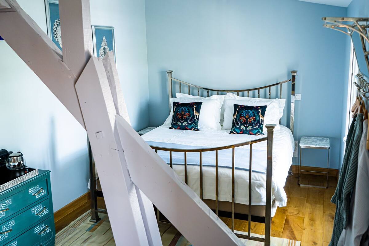 A bedroom with a metal bed frame, white bedding, and decorative pillows. There is a small bedside table and a dresser with drawers.