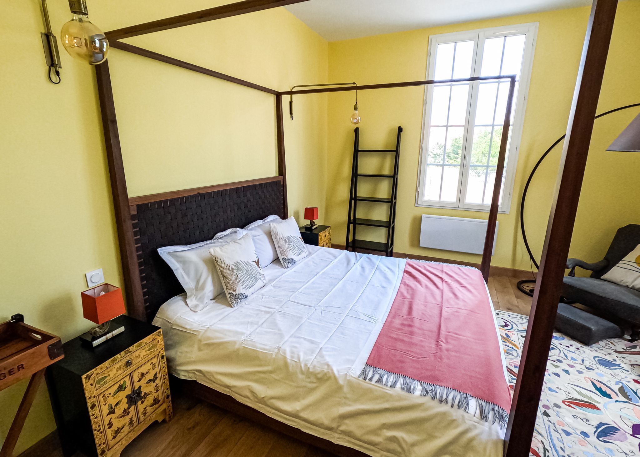 Un lit à baldaquin avec une structure en bois foncé et un couvre-lit blanc, une lampe rouge sur une table de chevet et une commode en bois.
