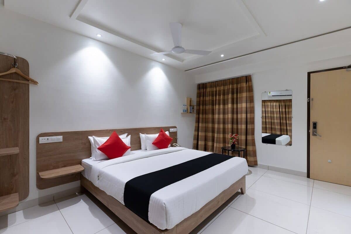 A modern hotel room with a wooden headboard, a large bed with white bedding and red pillows, a bedside table, a wardrobe, a ceiling fan, and a window with curtains.