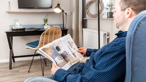 En mand læser et magasin i en moderne stue med et sort skrivebord, en fletstol og et fladskærms-tv på væggen.