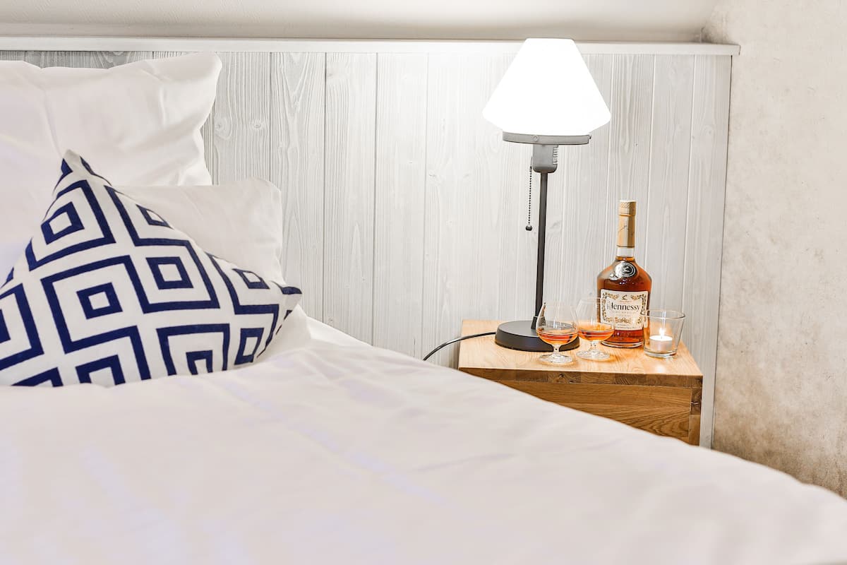 Ein ordentlich bezogenes Bett mit einem gemusterten Kopfkissen, einer Nachttischlampe, einer Whiskeyflasche und zwei Gläsern auf einem Holznachttisch.