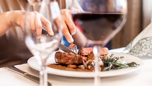 En person skærer kød på en tallerken med kniv og gaffel, og et glas rødvin.