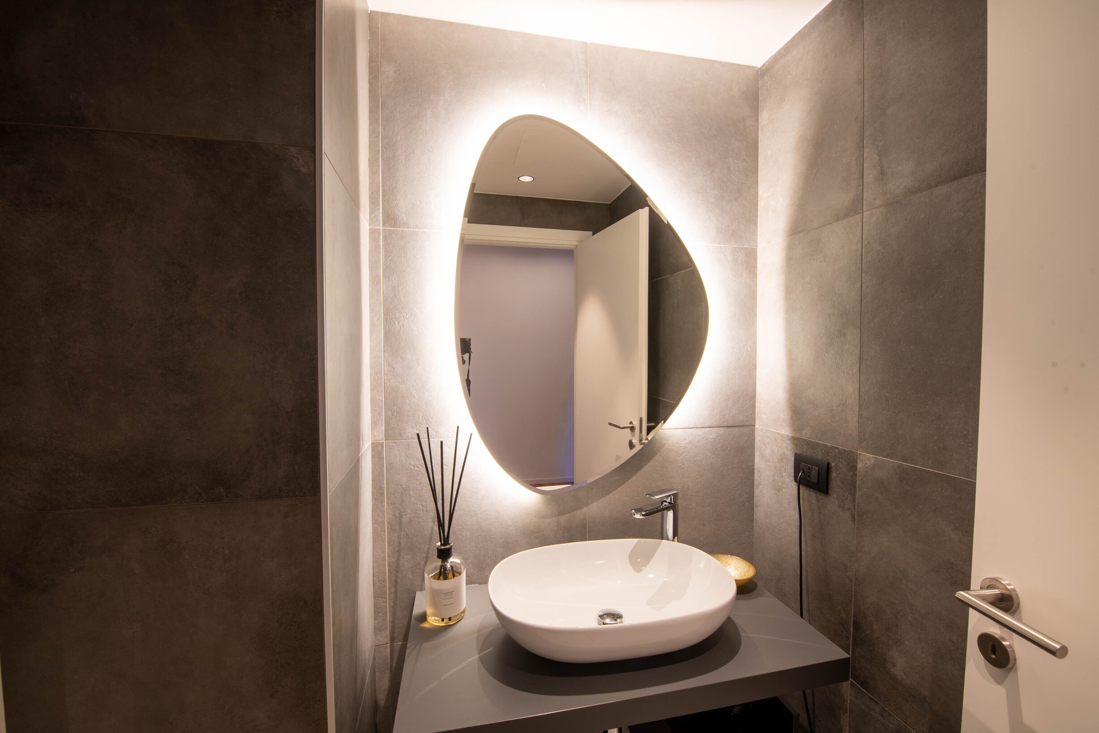 A modern bathroom with a large oval mirror, a white sink, and a small bottle of reed diffuser.