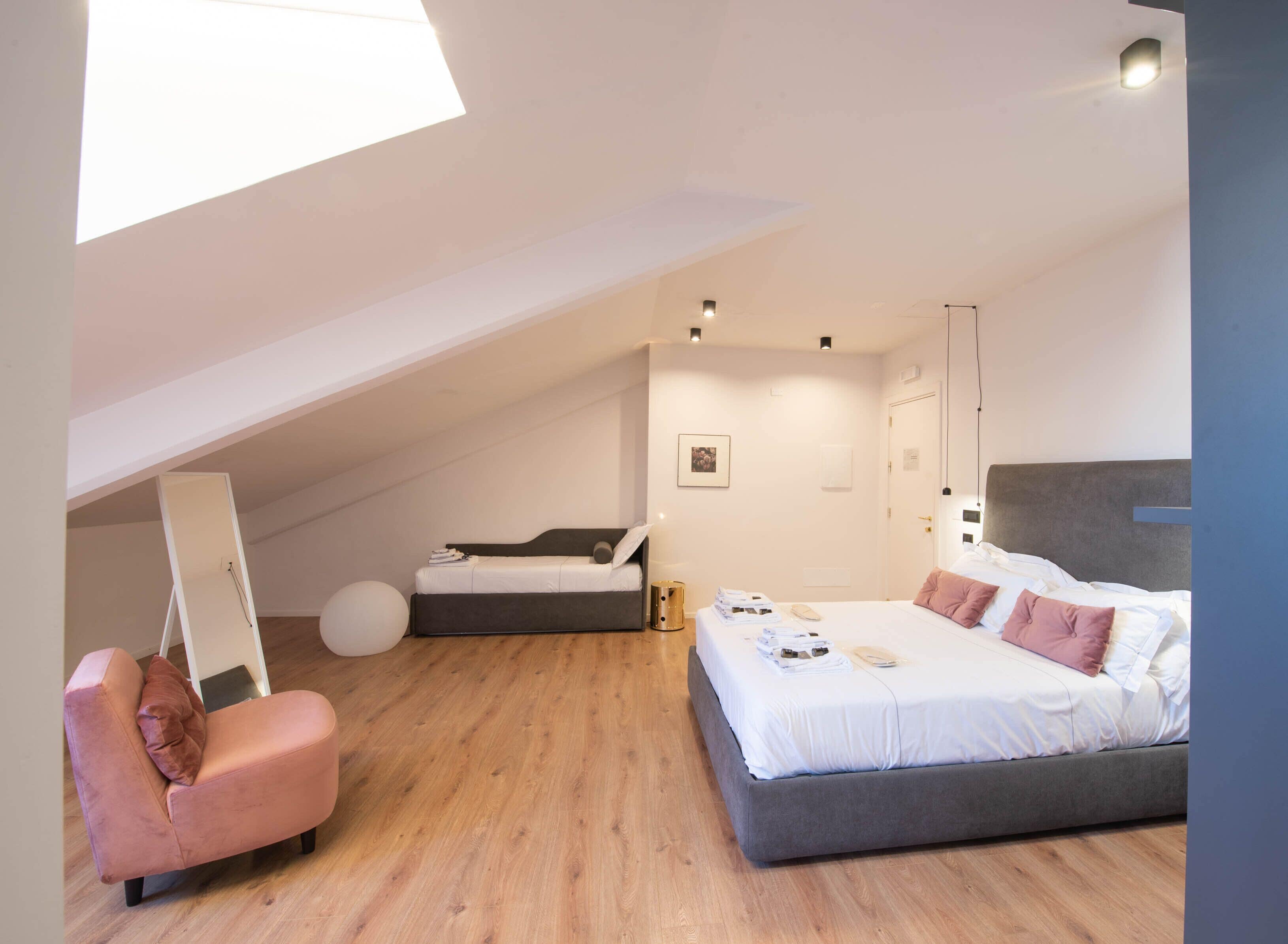Een slaapkamer met een loft-plafond, een bed met veel kussens, een klein nachtkastje en een roze stoel.