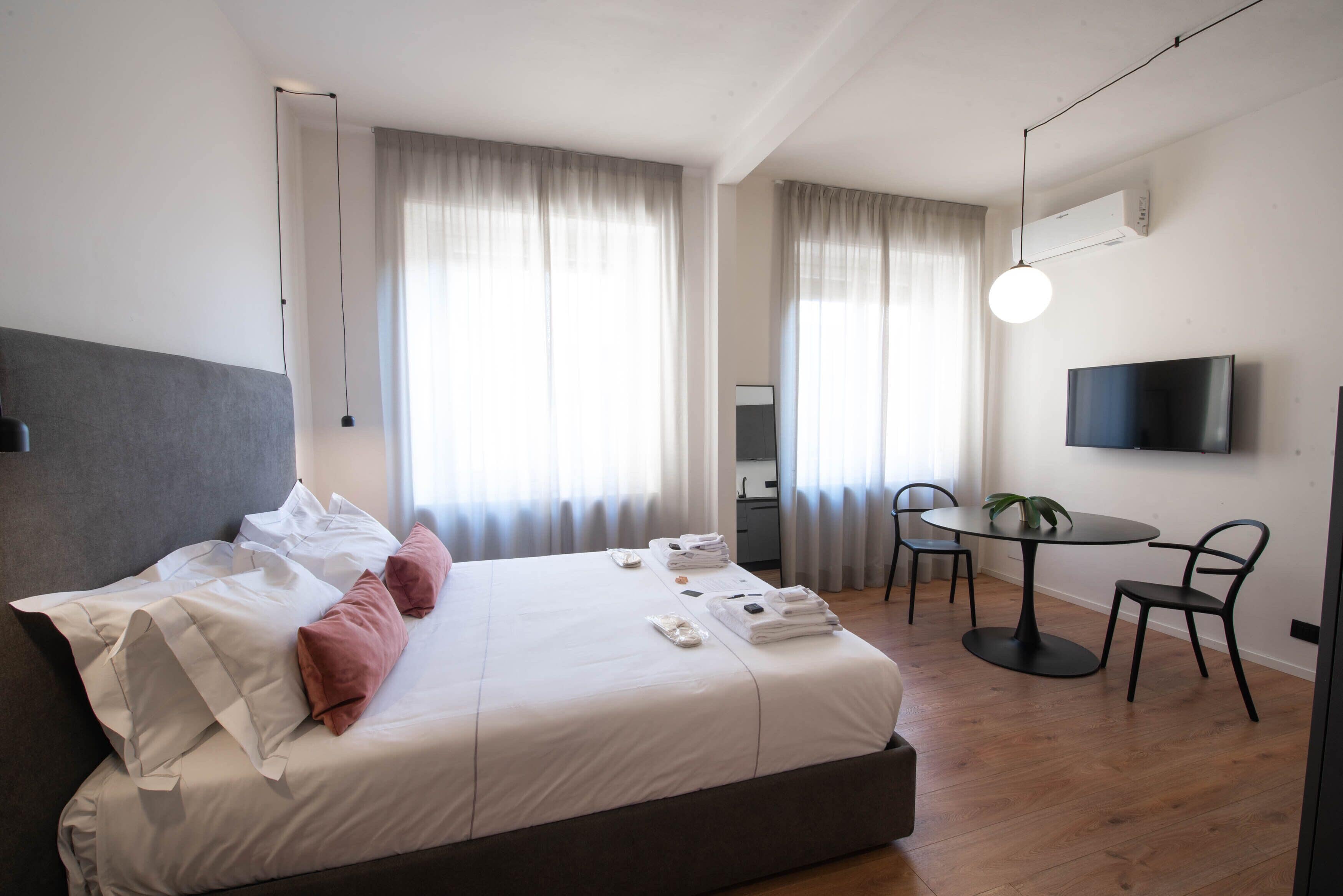 A modern hotel room with a large bed, a round dining table, two chairs, a TV, and a potted plant.