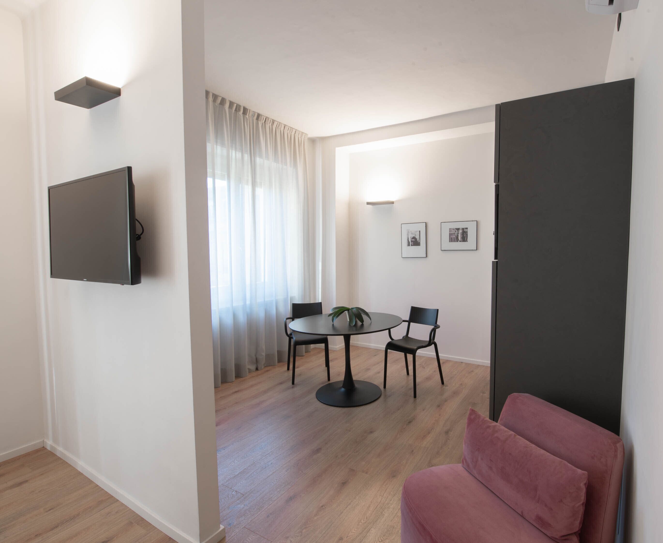 Un salon moderne avec un téléviseur à écran plat fixé au mur, une table de salle à manger ronde avec des chaises, un canapé rose et une grande fenêtre ornée de rideaux légers.