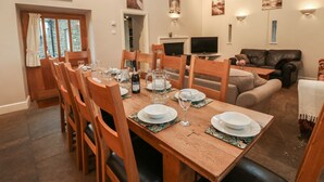 Un coin repas avec une table en bois dressée pour six personnes, entourée de chaises. Un téléviseur est fixé au mur, et un canapé est visible au fond.