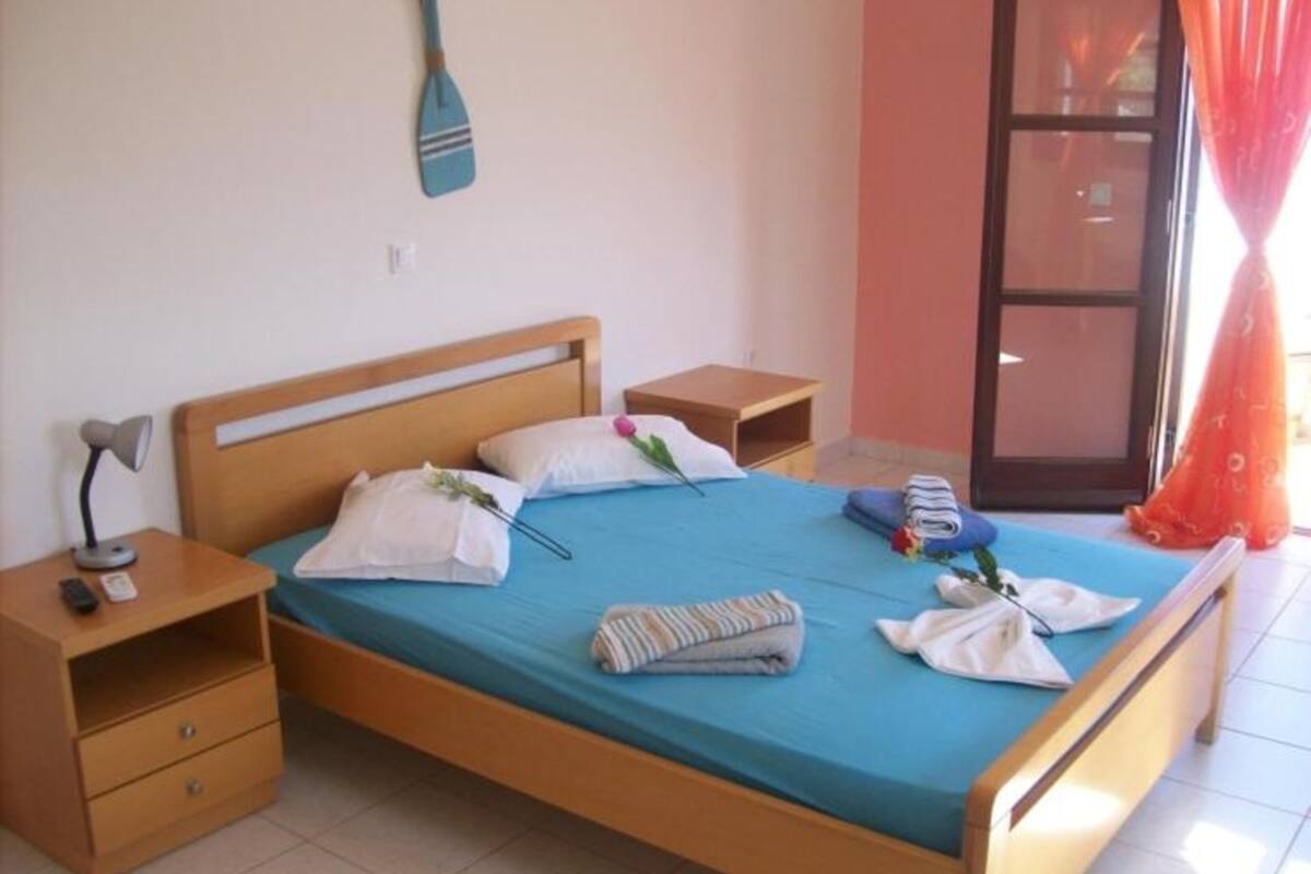 Un dormitorio con una cama de madera, dos mesitas de noche, un velador y una ventana con cortinas, además de un objeto fijado a la pared.