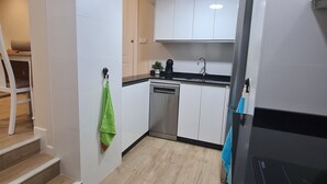 Una cocina moderna con armarios blancos, lavavajillas y una estufa negra. Se ve una escalera de madera que sube a otra habitación.