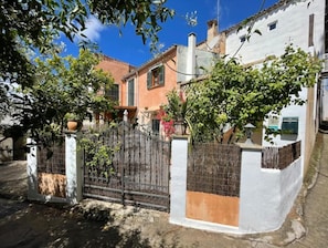 Uma área residencial com portão de entrada, um prédio branco com telhado vermelho e uma árvore em primeiro plano.
