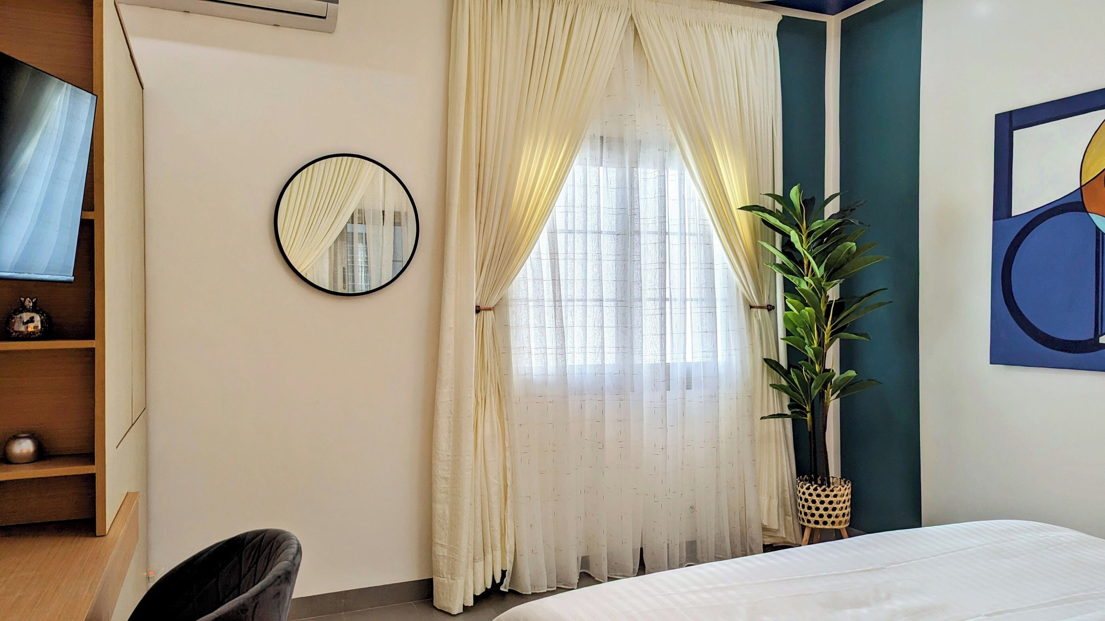 Un dormitorio con cama, un espejo redondo, una ventana con cortinas traslúcidas, una planta en maceta y un cuadro abstracto en la pared.