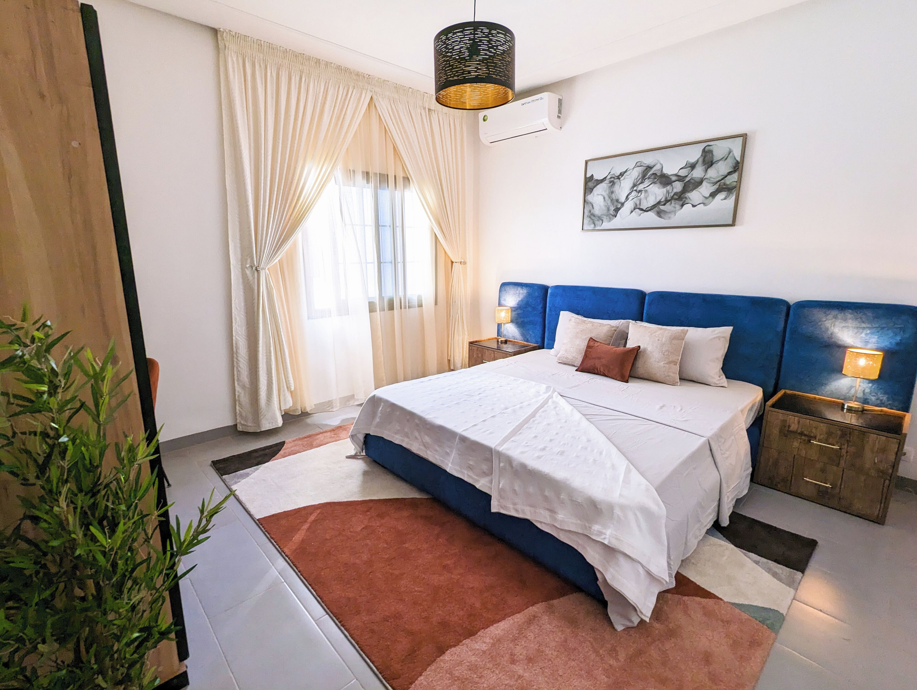 A bedroom with a large bed, a nightstand with a lamp, a wall-mounted air conditioner, and a window with sheer curtains.