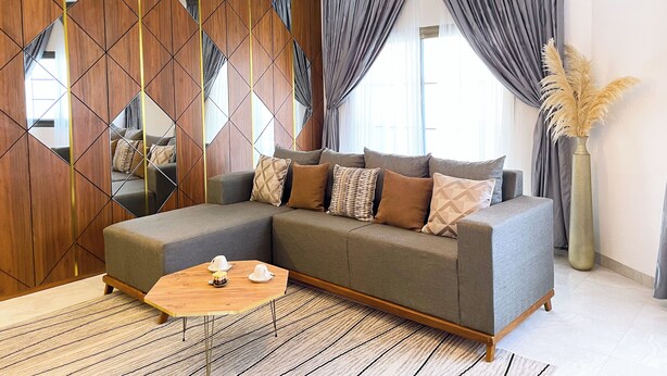 Un salon moderne avec un canapé d’angle, une cloison en bois ornée de motifs géométriques, un grand miroir et un vase contenant des plumes d’herbe des pampas.