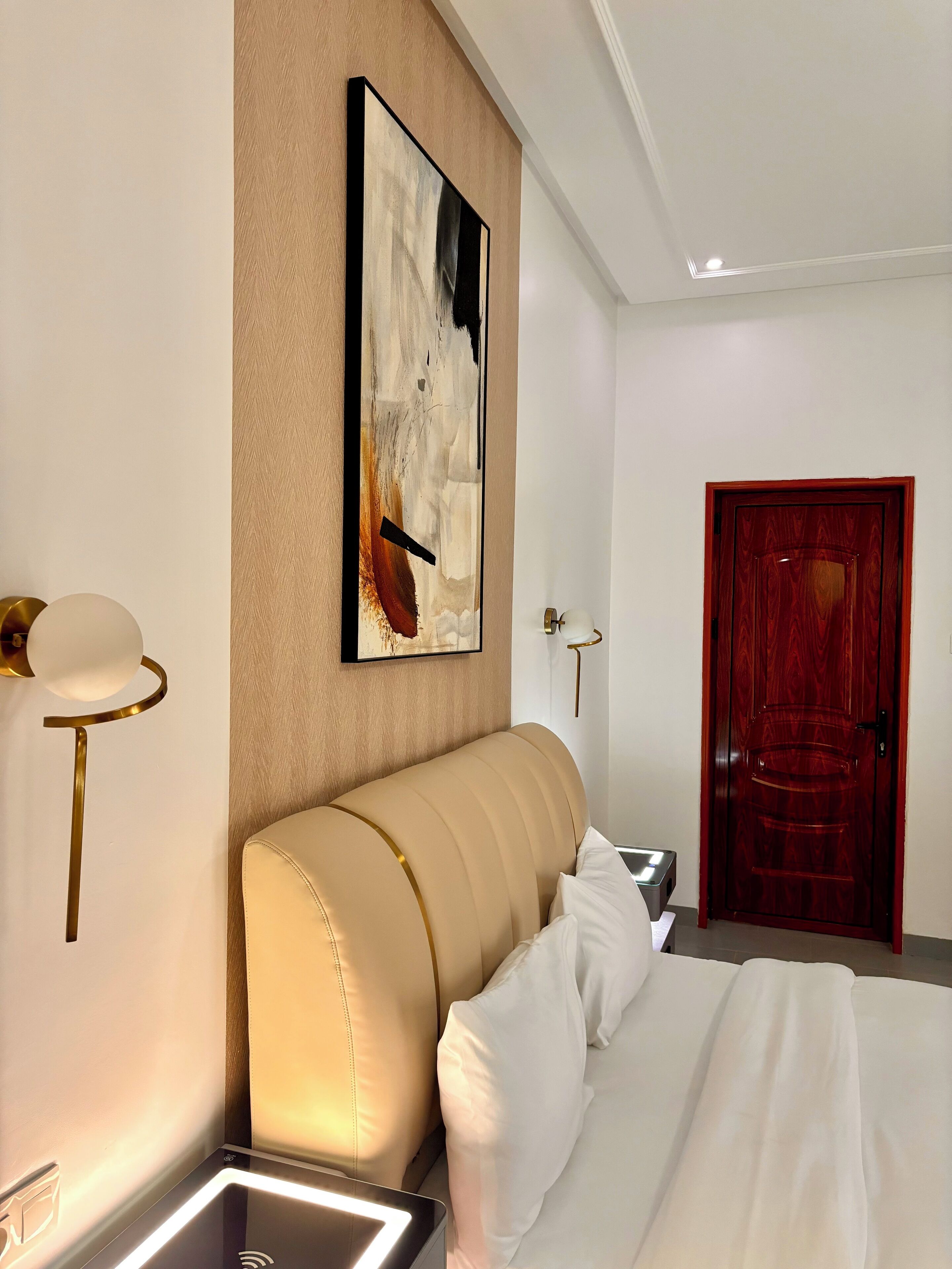 A hotel room with a bed, a painting on the wall, a door, and a wall-mounted light fixture.