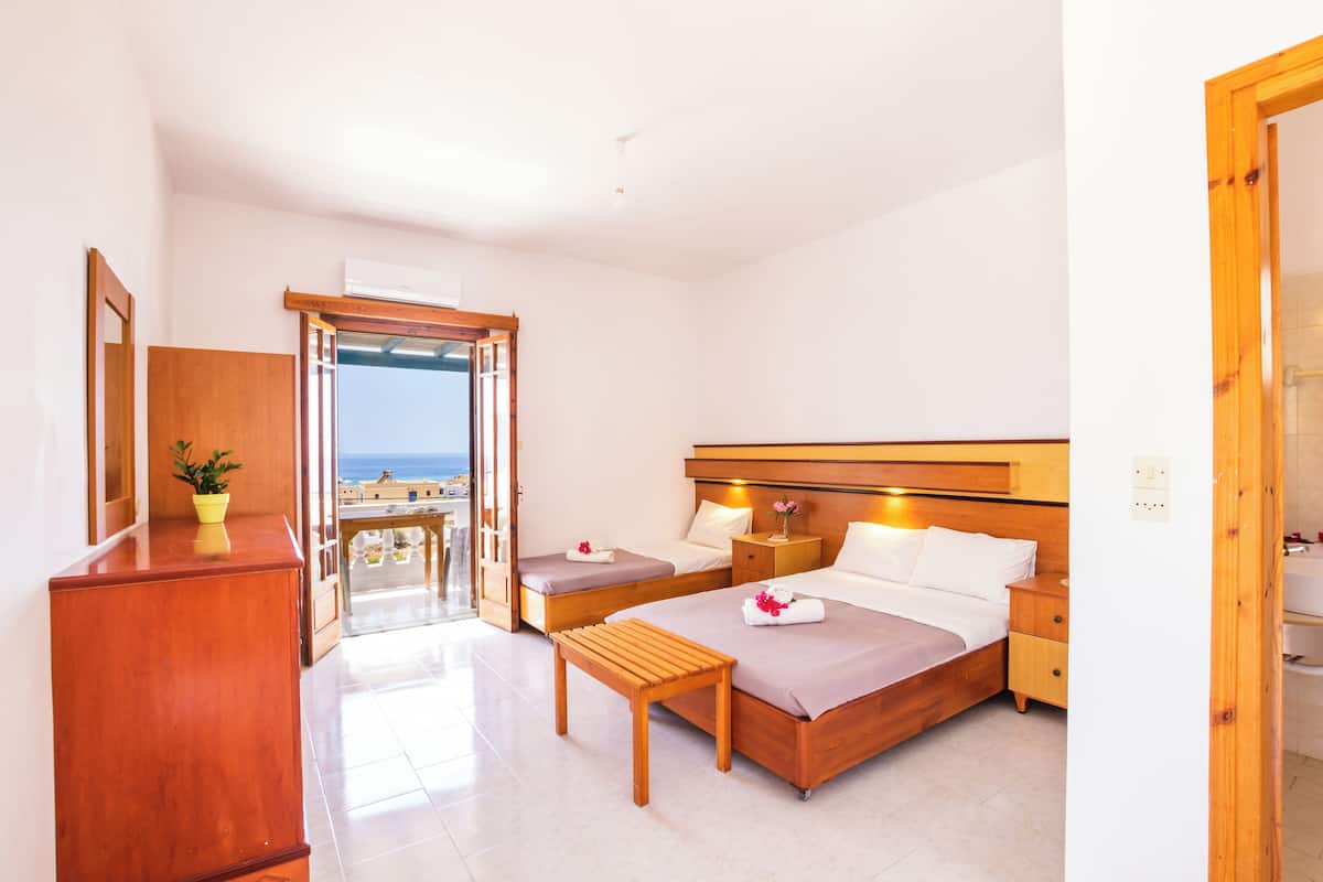 Una habitación de hotel con una cama, una mesita de noche y un pequeño banco de madera. Hay una ventana con vista al mar.