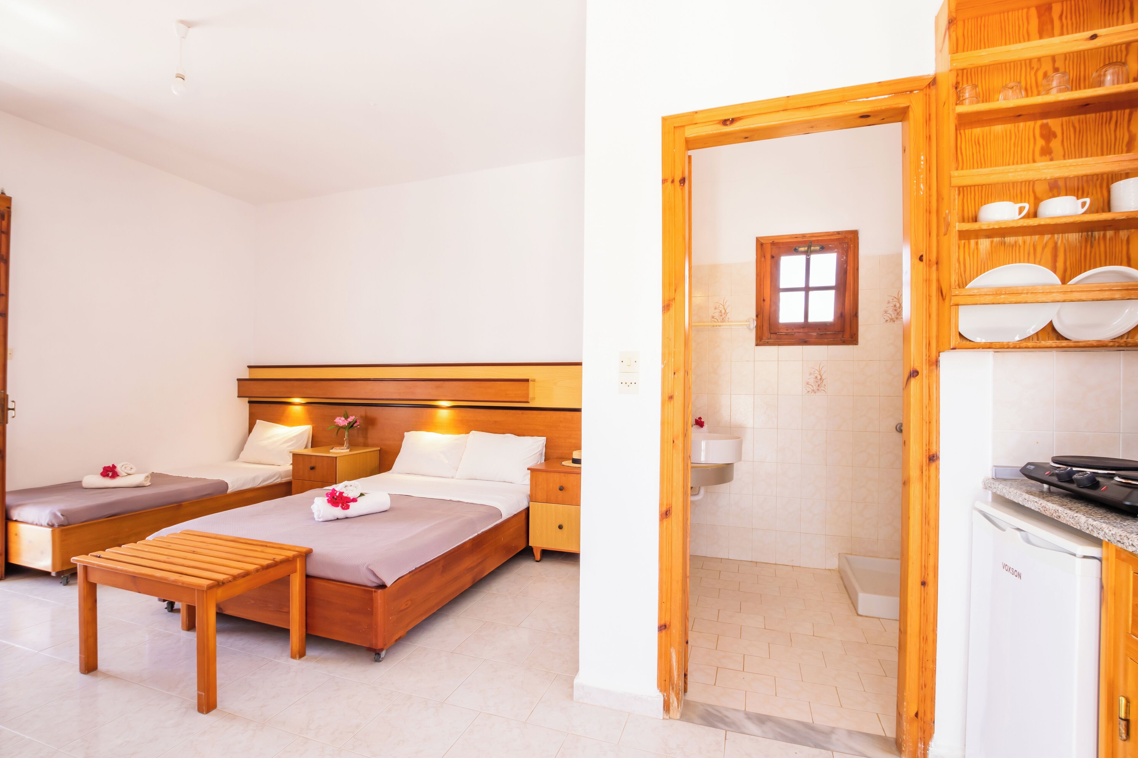 Habitación de hotel con dos camas, una mesa de madera, una mesita de noche pequeña y una kitchenette con un lavamanos y estufa.