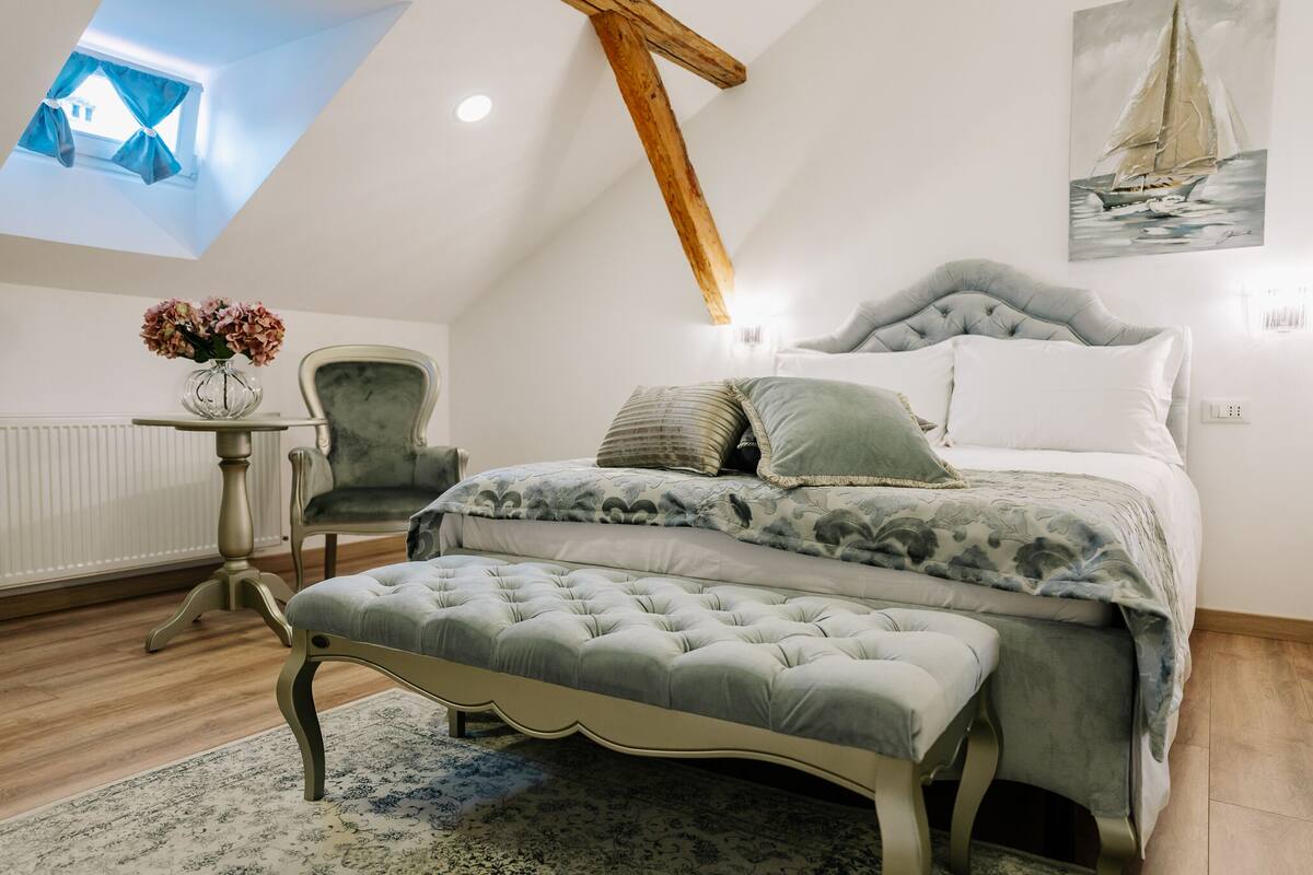 A bedroom with a bed, a chair, a table with a vase, and a painting on the wall.