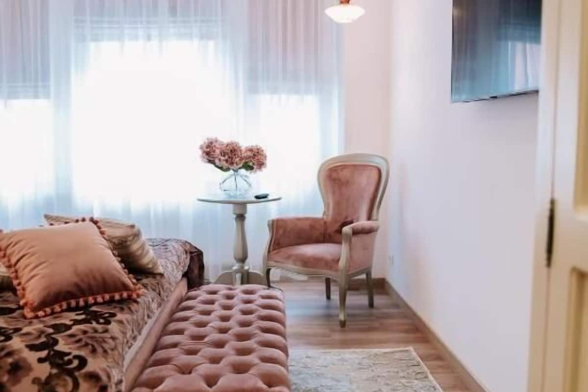 A bedroom with a bed, a bench, a chair, a small table with flowers, and a TV on the wall.