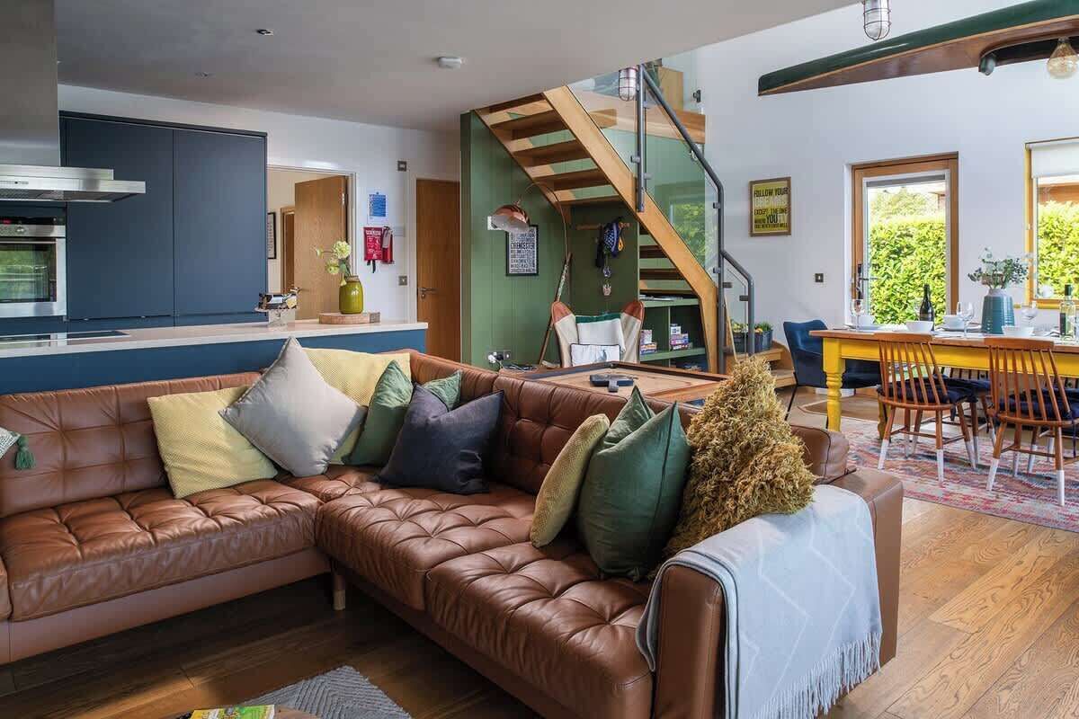 A modern living room with a brown leather sofa, a dining area with yellow chairs, and a staircase leading to an upper level.