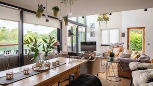 A modern living room with a wooden dining table, comfortable seating, and large windows offering a view of greenery.
