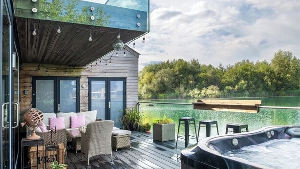A modern outdoor deck with a glass enclosure, wooden deck, seating area with wicker furniture, and a hot tub.
