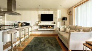 A modern kitchen with a built-in stove, bar stools, and a flat-screen TV mounted above the fireplace. - Grebe Lodge (South Cerney)