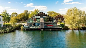 A modern houseboat with a glass facade, situated on a calm river. - Grebe Lodge (South Cerney)