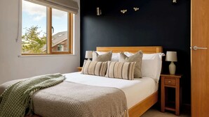 A bedroom with a large window, a bed with multiple pillows, bedside tables with lamps, and a dark wall with decorative elements. - Grebe Lodge (South Cerney)