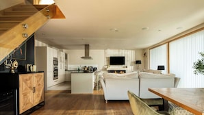 A modern kitchen with a wooden staircase, a central island, and a dining area with a wooden table and chairs. - Grebe Lodge (South Cerney)