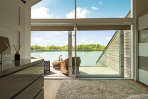 A modern interior with a large sliding glass door leading to a balcony with a view of a lake and trees. - Malachite Lodge (South Cerney)