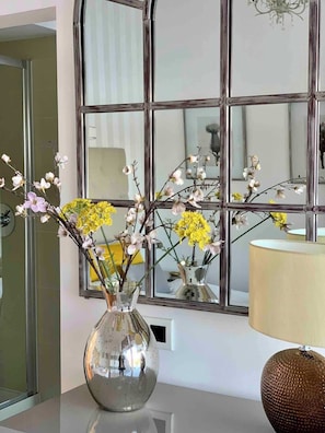 A room with a large mirror, a vase with flowers, and a lamp.