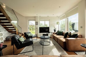 A modern living room with a staircase, a flat-screen TV, and large windows. - Malachite Lodge (South Cerney)