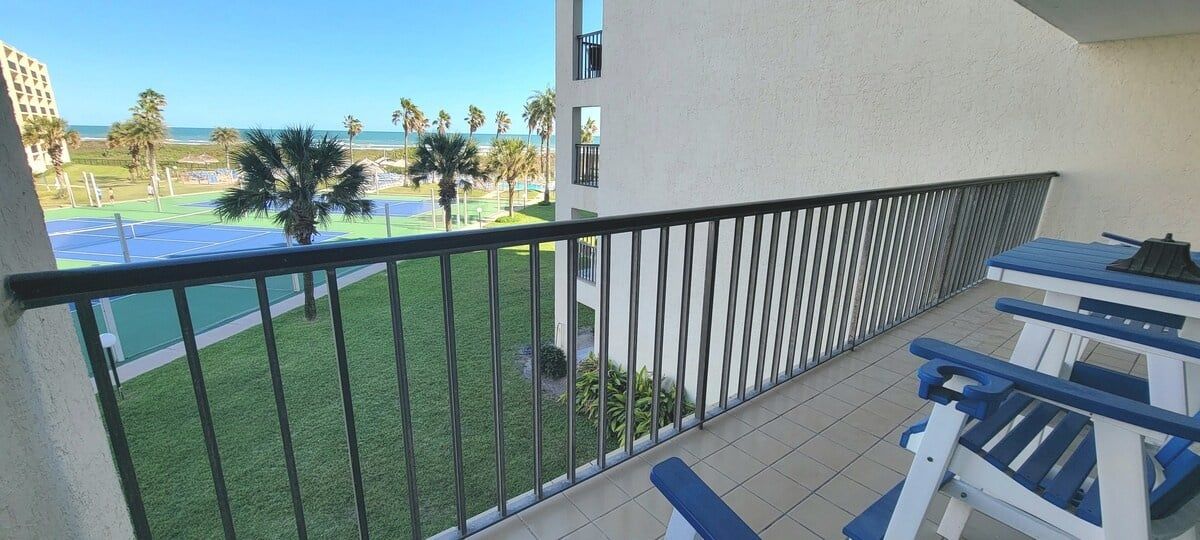 Un balcon donnant sur un court de tennis, des palmiers et un bâtiment en arrière-plan.