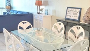 Un comedor con mesa de vidrio rodeada de sillas blancas, una lámpara colgante arriba y un sofá azul al fondo.