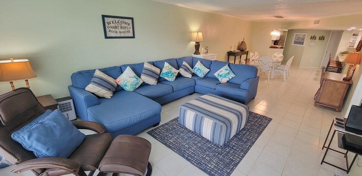 Una sala de estar con un sofá seccional azul, un sillón de cuero marrón, un otoman estampado y una mesita de noche con una lámpara.